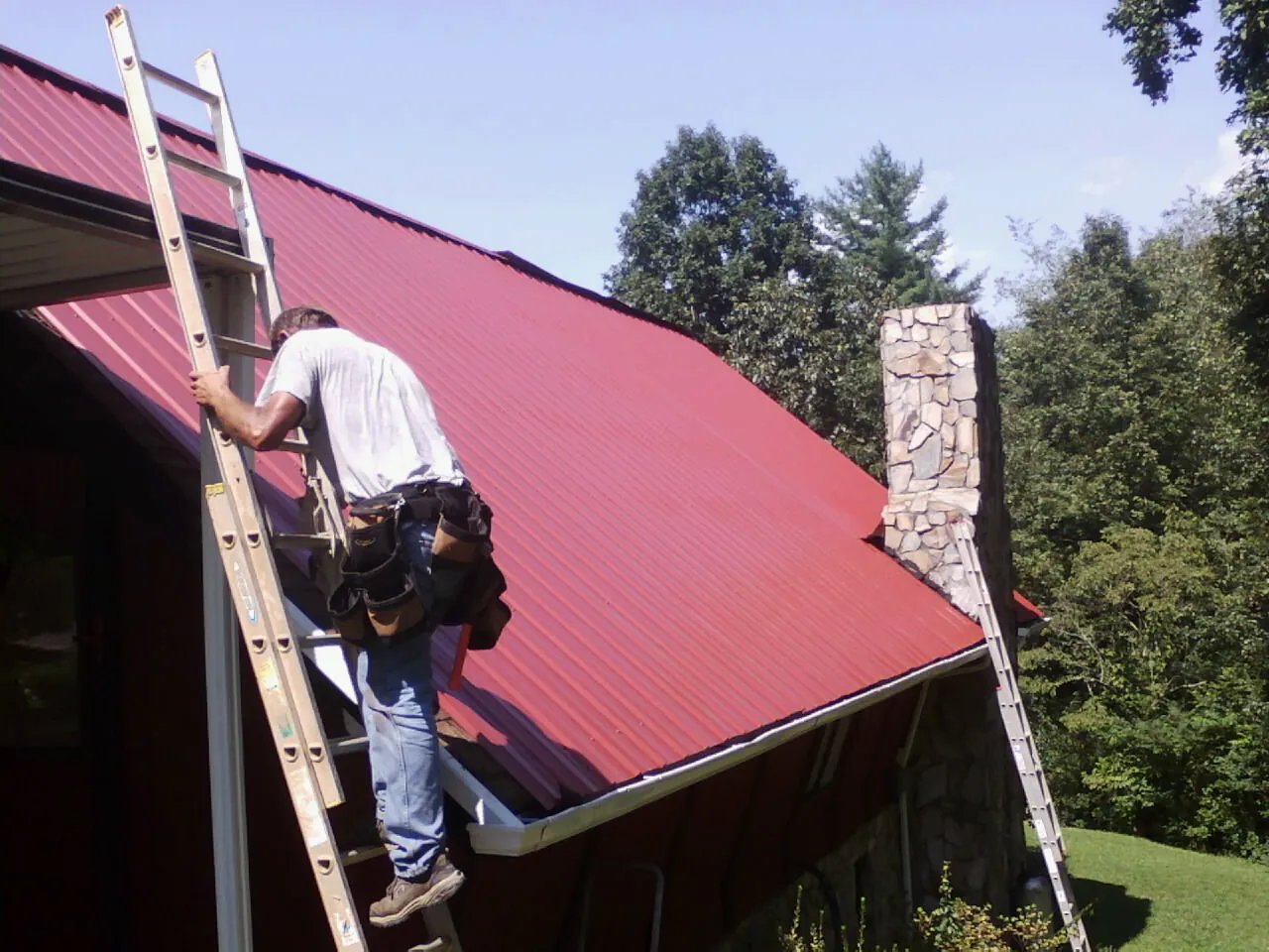 Roofer on a ladder beside a red metal roof and stone chimney — Soffit & Fascia Repair by Roofing Brothers in Emerald Isle